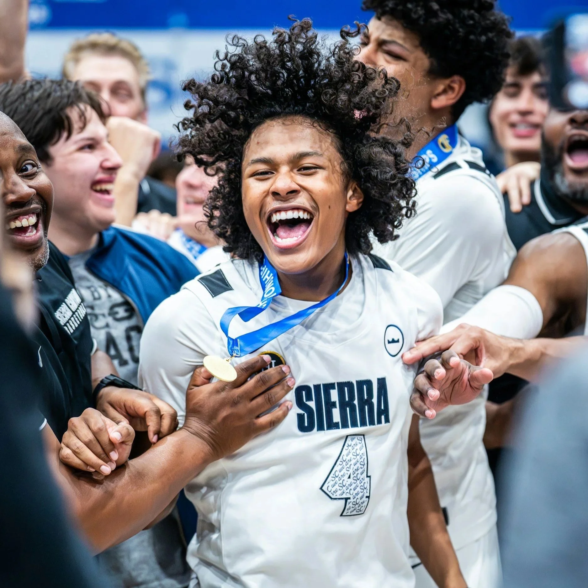A group of diverse young people celebrating, smiling, and holding hands, with one person wearing a medal and a shirt reading "Piedra Sierra."