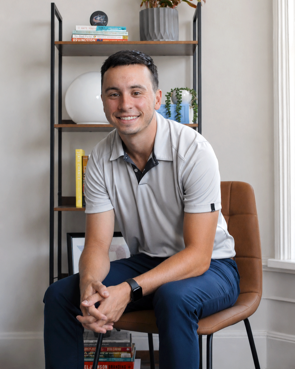A young man with dark hair, wearing a light gray polo shirt and dark blue pants, sits on a brown chair, smiling at the camera. Behind him is a black metal and wood bookshelf with decorative items, books, and plants, in a well-lit room with natural light from a window on the right.