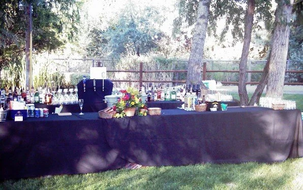 Outdoor table setup with drinks, glassware, and a flower arrangement in a park or backyard.