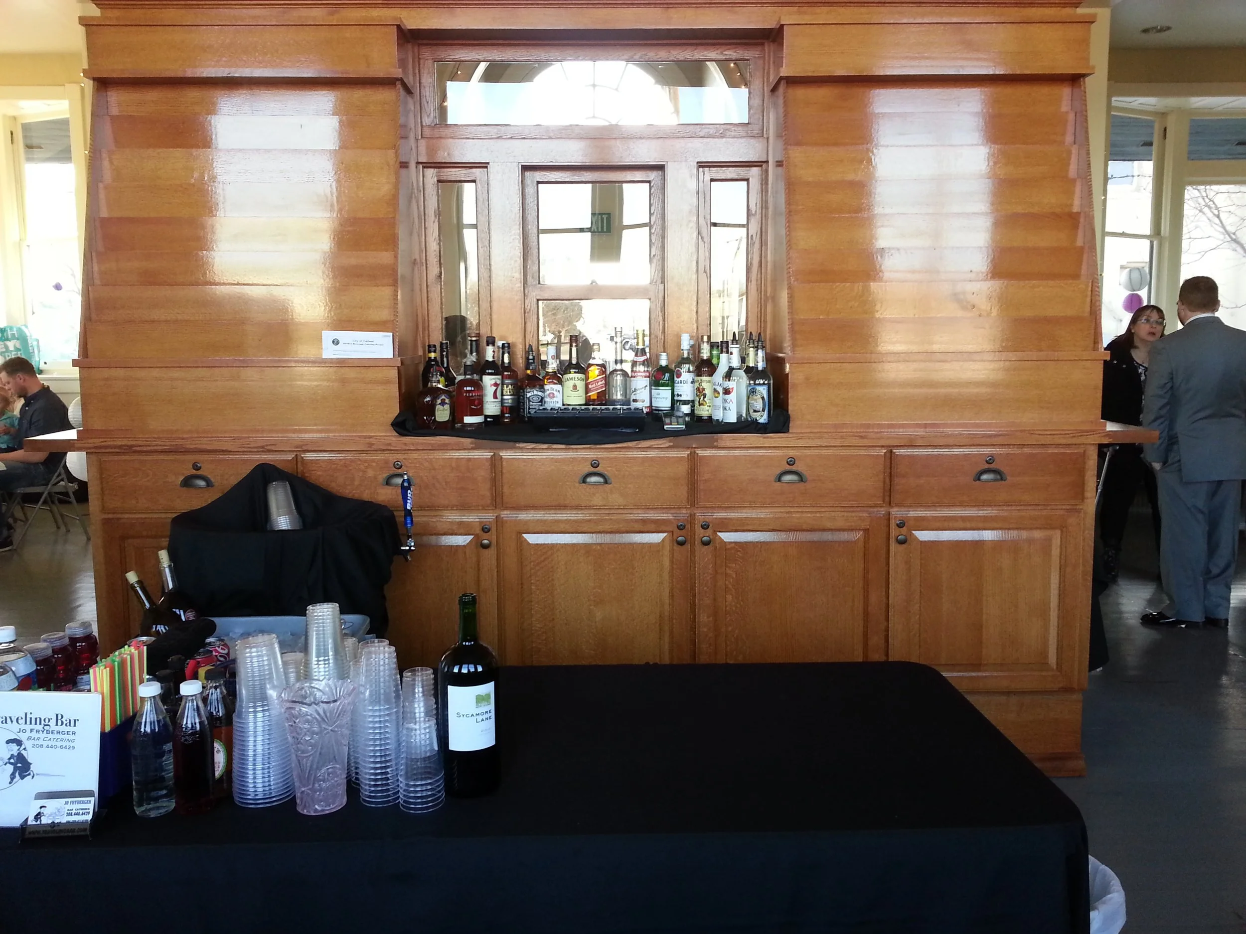 A wooden bar setup with various bottles of alcohol and cups on a black tablecloth. People are seen in the background at what appears to be an event or gathering.