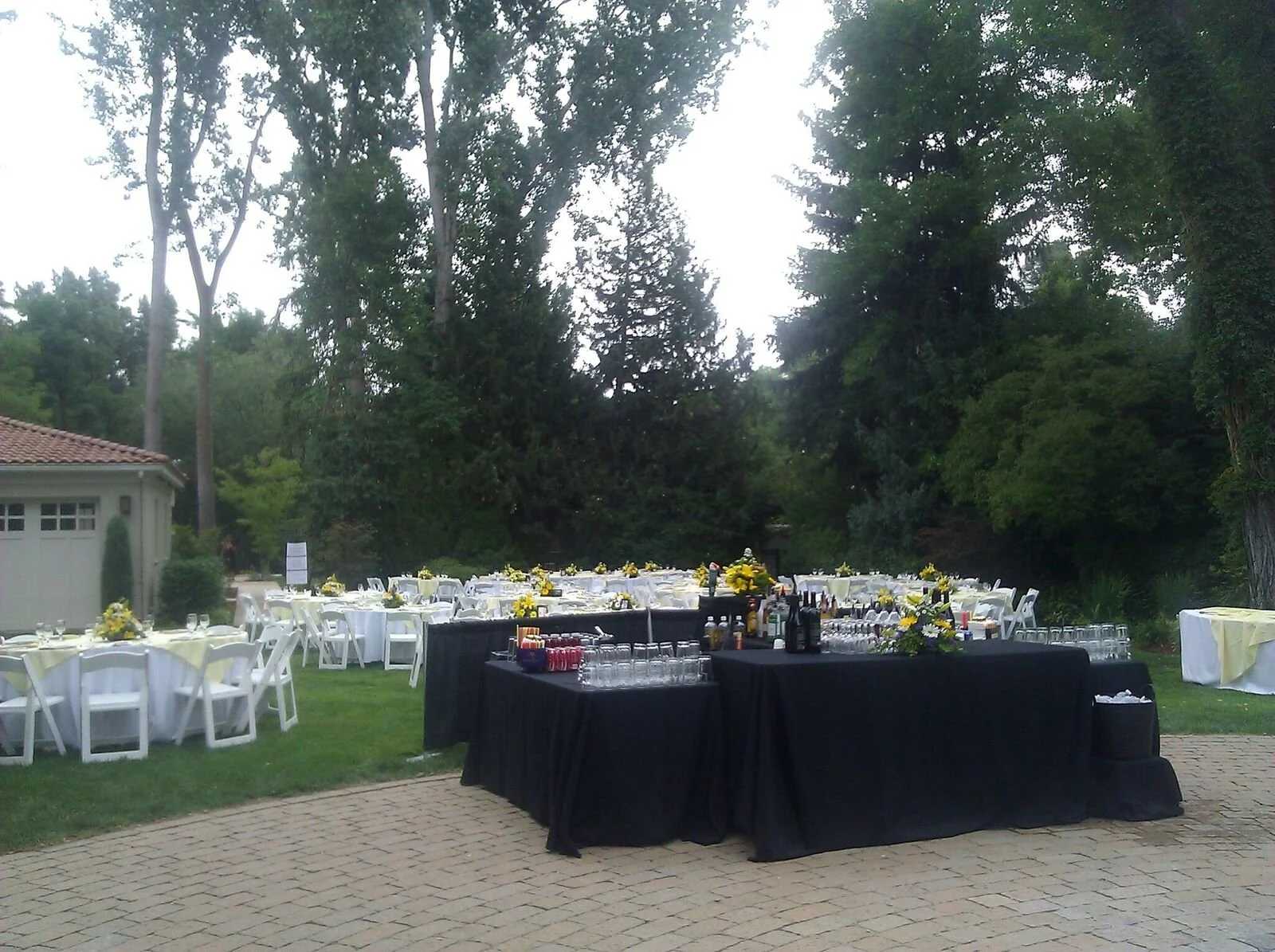 Outdoor wedding reception setup with round tables covered in yellow and white tablecloths, decorated with yellow flower centerpieces, situated on a grassy area surrounded by tall trees, with a black catering table in the foreground and a small buildi