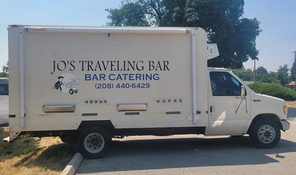 White delivery truck with a sign reading 'Jo's Traveling Bar, Bar Catering' and a phone number, parked in a lot with trees in the background.