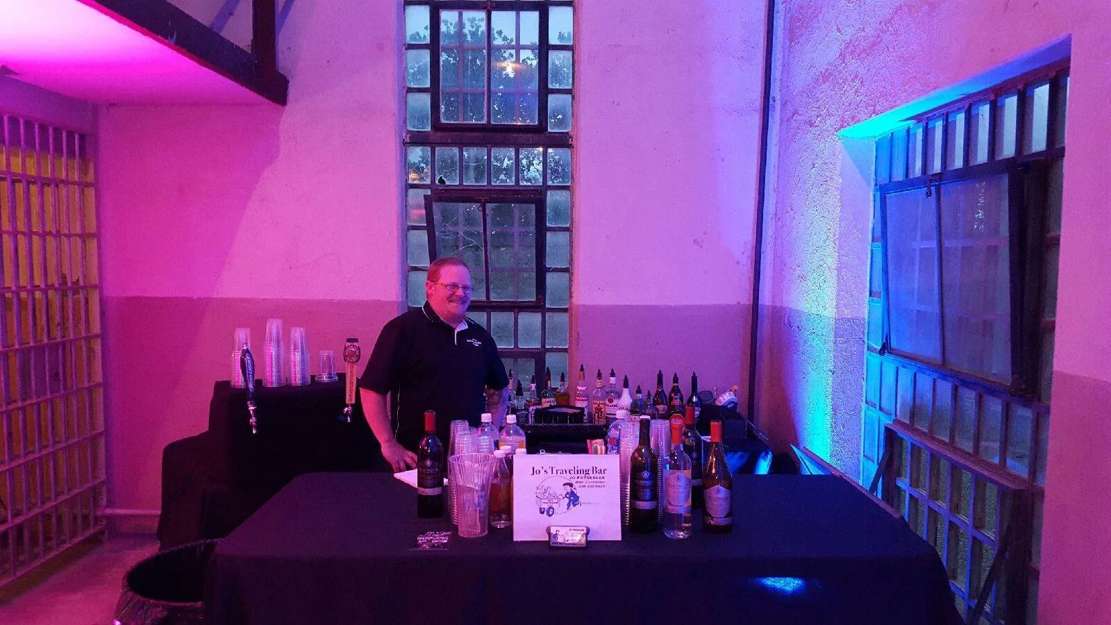 Bartender standing behind a bar with various bottles in a dimly lit room illuminated by pink and blue lights.