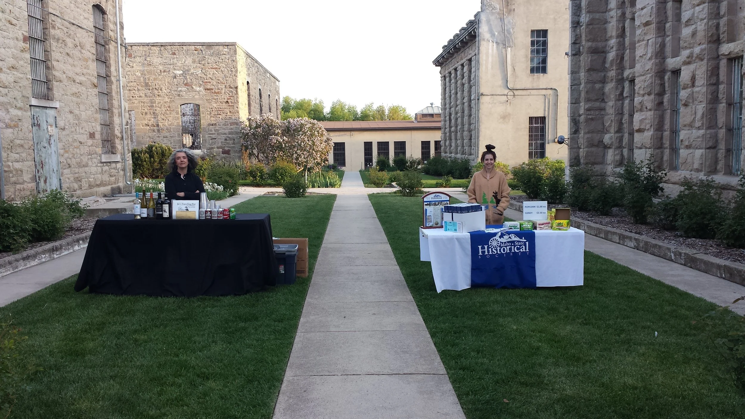 Two women with tables set up for an event or sales, standing on a sidewalk between historic stone buildings with a garden in the background.