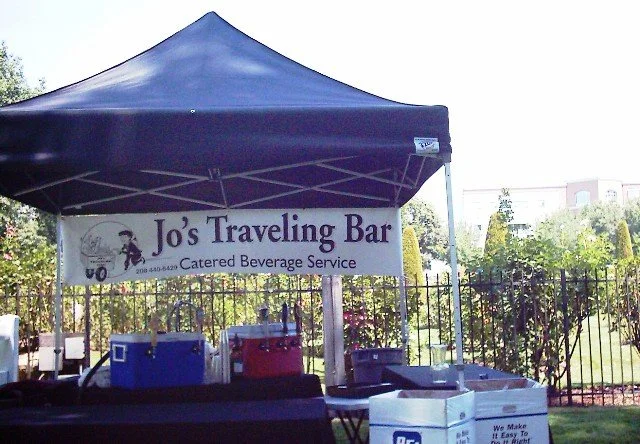 Outdoor setup with a blue canopy tent and a banner for Jo's Traveling Bar and Catering Beverage Service. Coolers and supplies are underneath the tent, with a fence, trees, and buildings in the background.