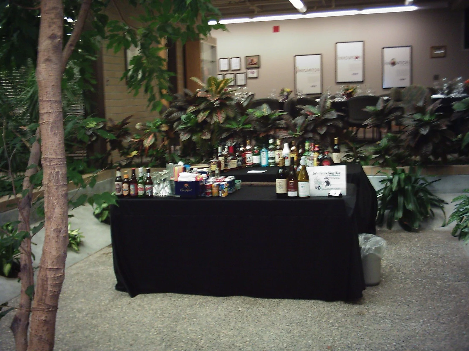A table with various bottles of alcohol and drink mixers, set up for a bar at an indoor event in a venue with plants and framed posters or menus on the wall.