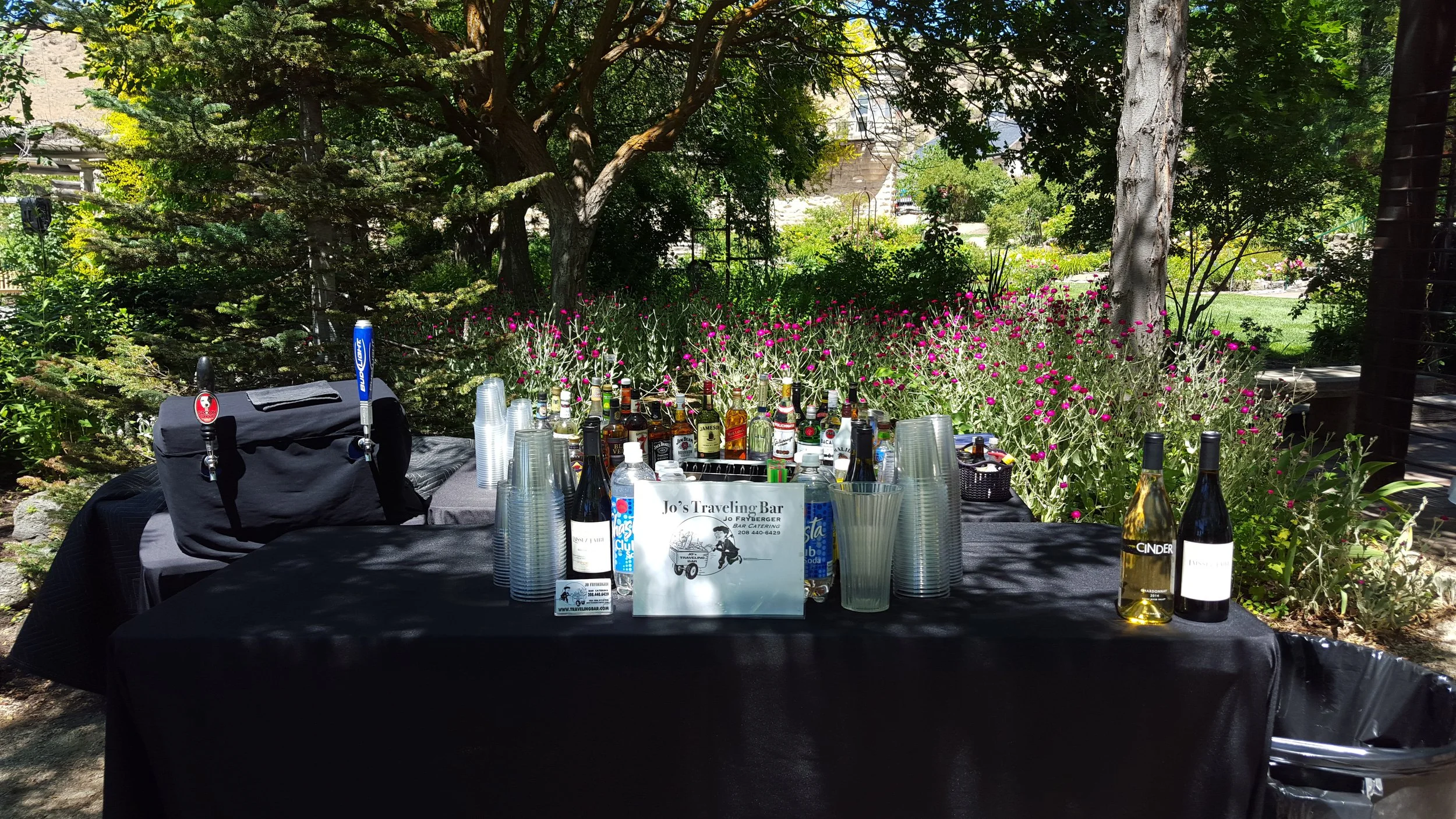 Outdoor bar setup with various bottles of alcohol, empty plastic cups, and banners on a table in a garden with green trees and pink flowers in the background.