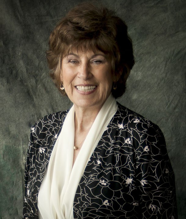A smiling older woman with short brown hair, wearing a black blazer with a white floral pattern and a white blouse, against a green textured background.