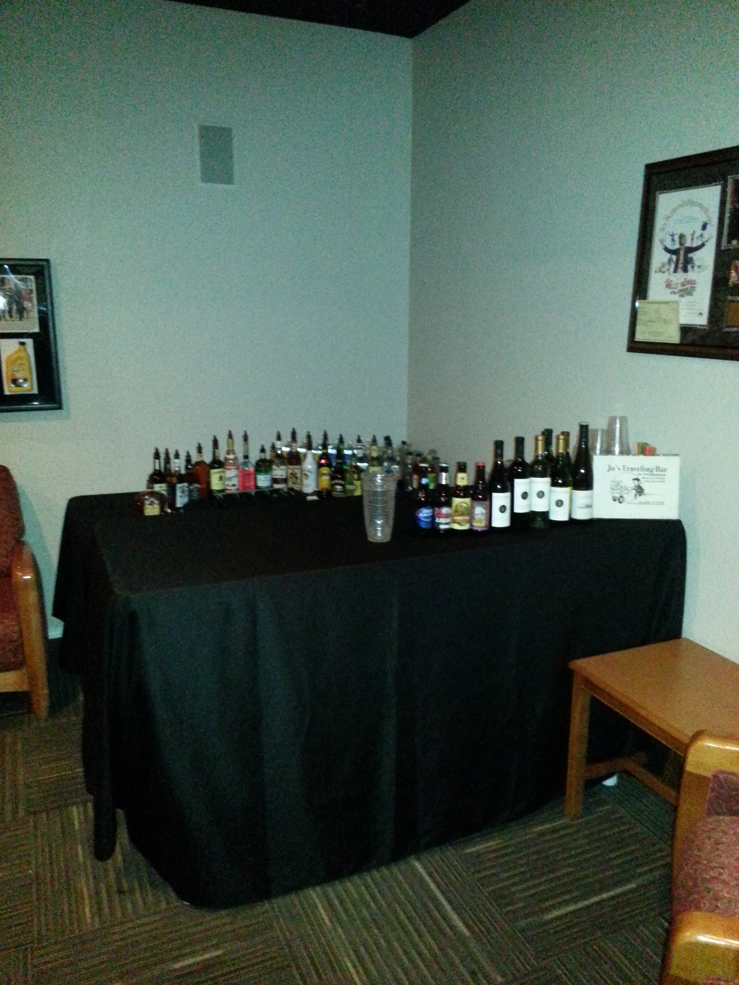 A table covered with a black tablecloth displaying various bottles of alcohol, with plastic cups and promotional material, set in a room with framed pictures on the wall and a striped carpet.
