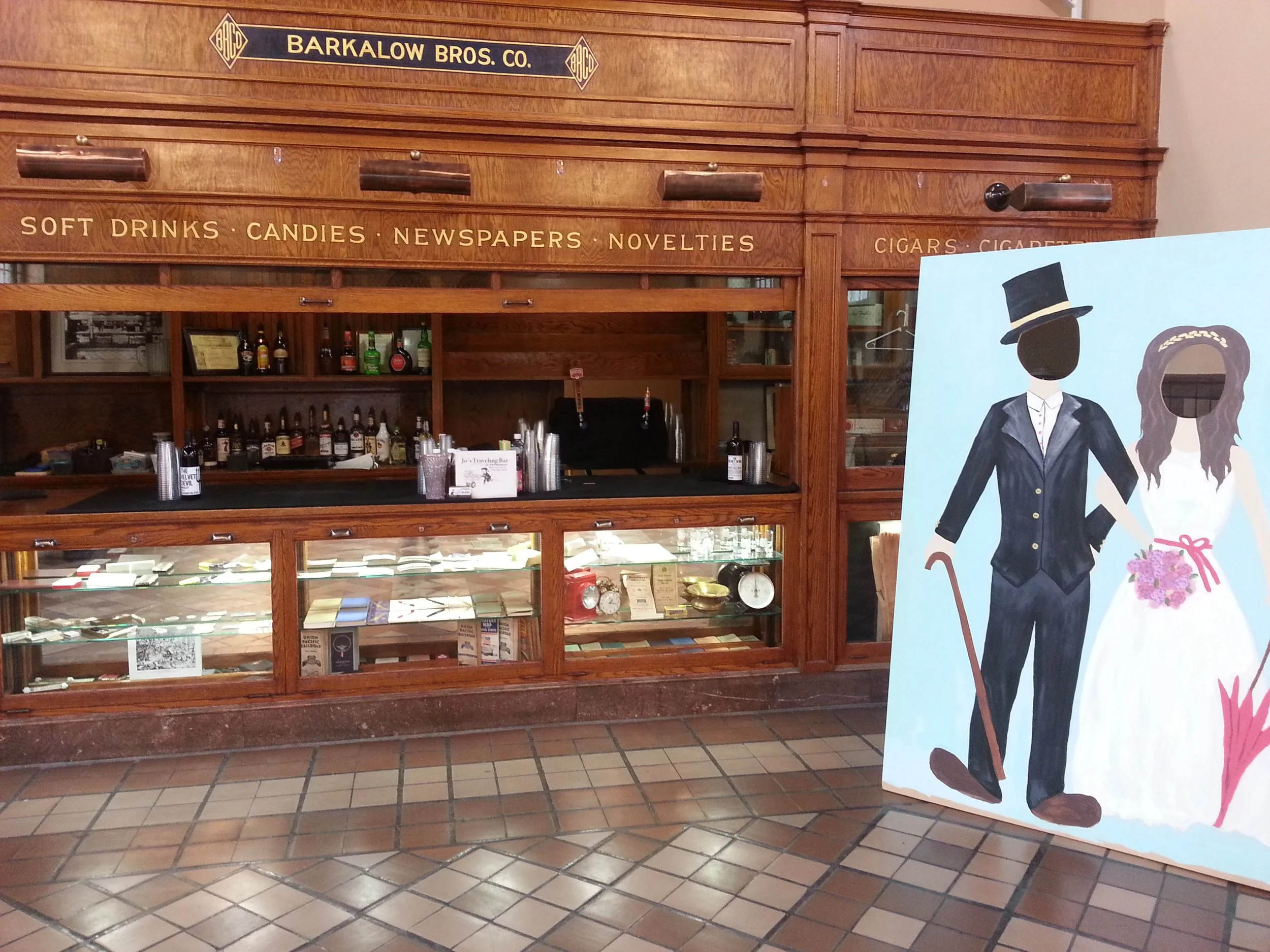 A wooden display case with bottles of alcohol and a framed certificate inside. Above, engraved on the wood, is the text 'BARKALOW BROS. CO.' and labels for items sold including 'SOFT DRINKS,' 'CANDIES,' 'NEWSPAPERS,' 'NOVELTIES,' 'CIGARS,' and 'CIGAR