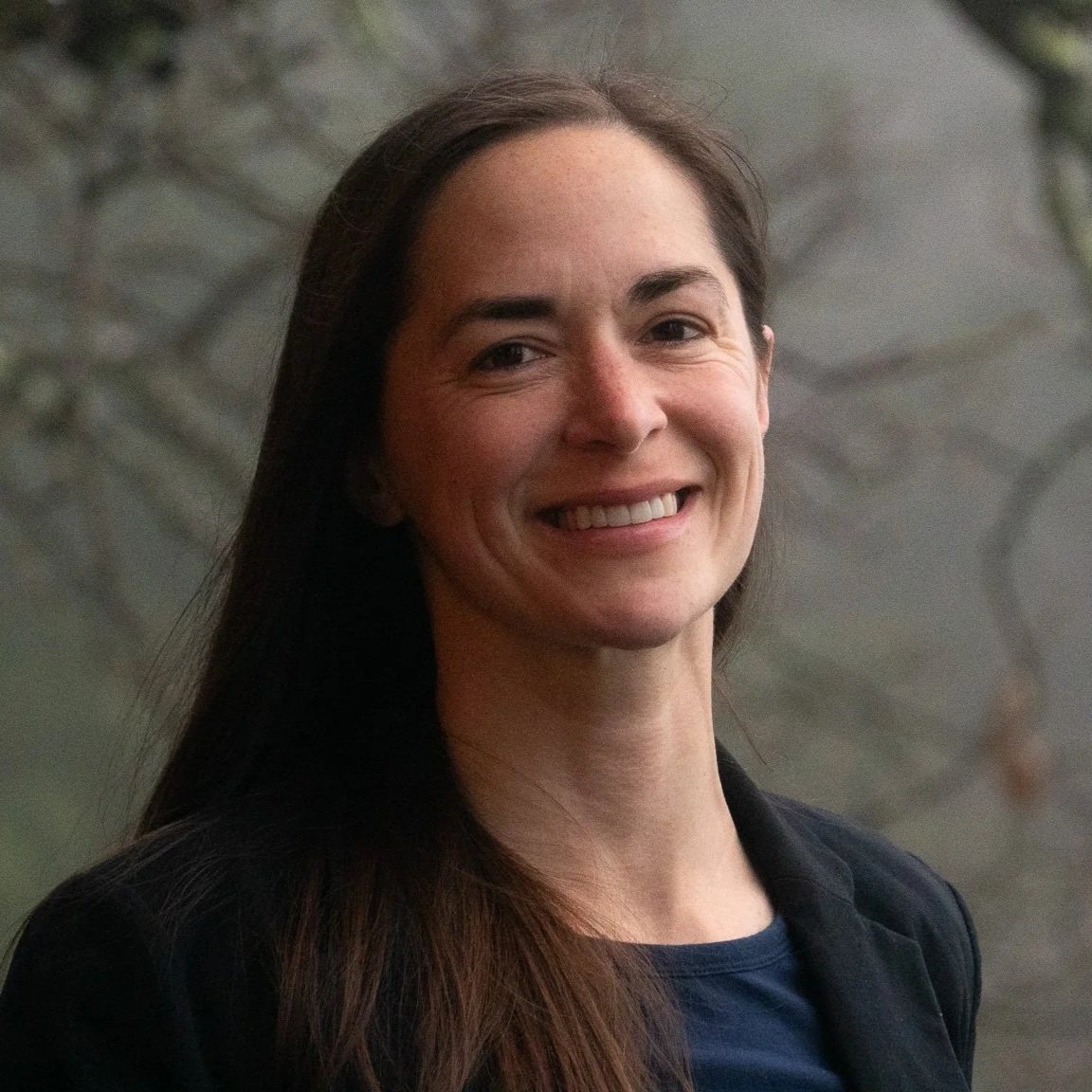 A woman smiling outdoors with trees in the background.