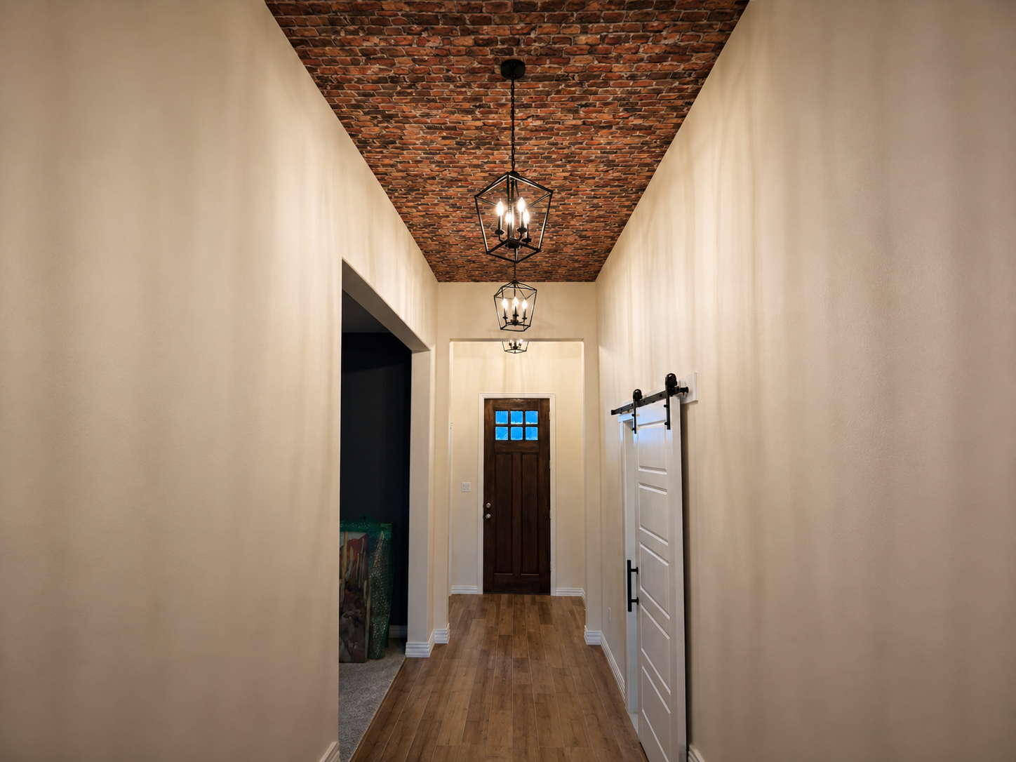 Hallway with white walls, a brick ceiling, wooden floor, and black lantern-style ceiling lights. There is a barn door on the right and a paneled wooden front door at the end with small window squares at the top.