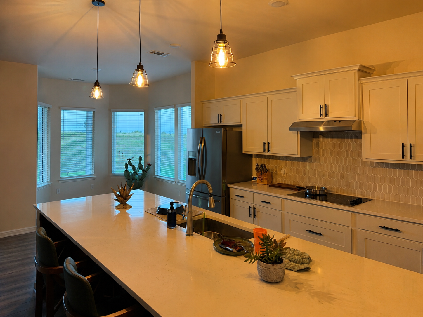 Modern kitchen with white cabinetry, stainless steel appliances, a large island with a sink, and pendant lights. Large windows in the background show a view of the outdoors with greenery.