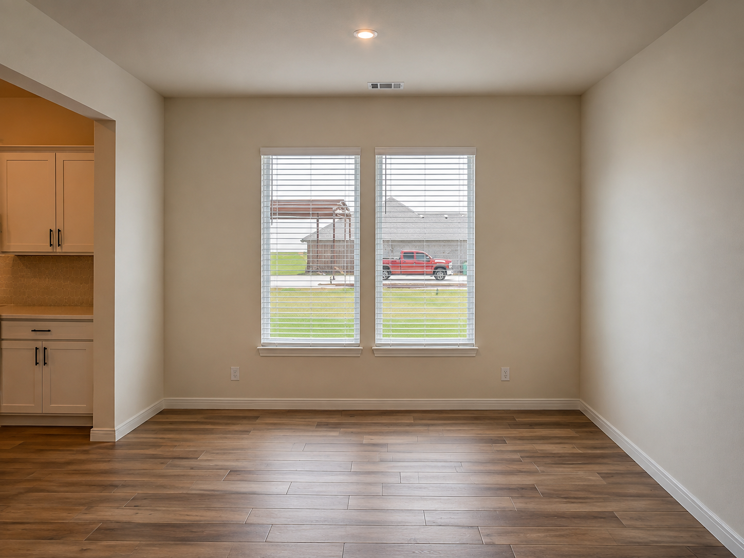 Empty room with hardwood flooring, two windows with blinds, view of a suburban street with a red truck and house outside.