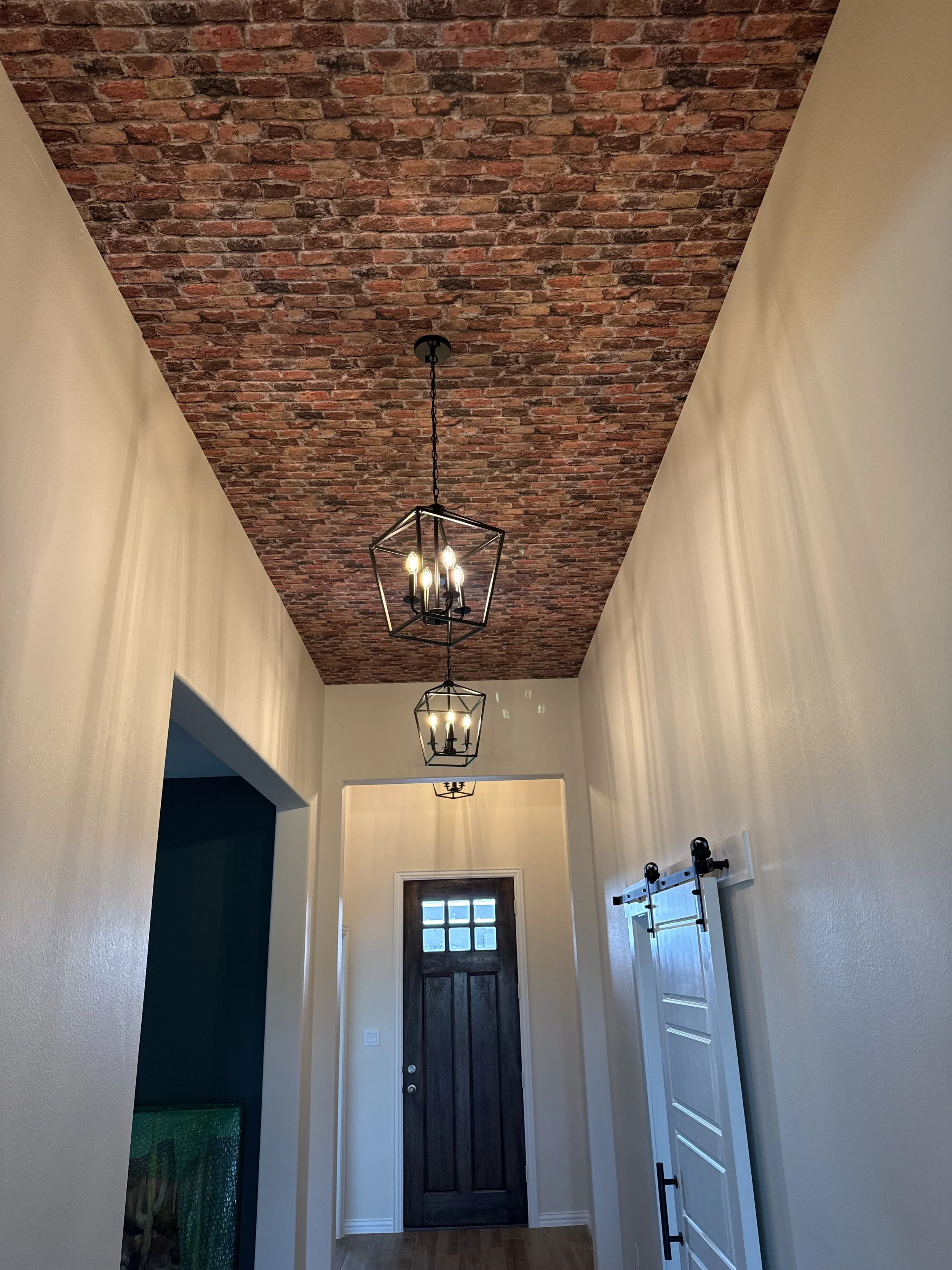 View toward the front door of a remodeled entry hallway showing new pendant lighting and brick-style ceiling wallpaper, a Dallas-Fort Worth renovation by MZL Construction