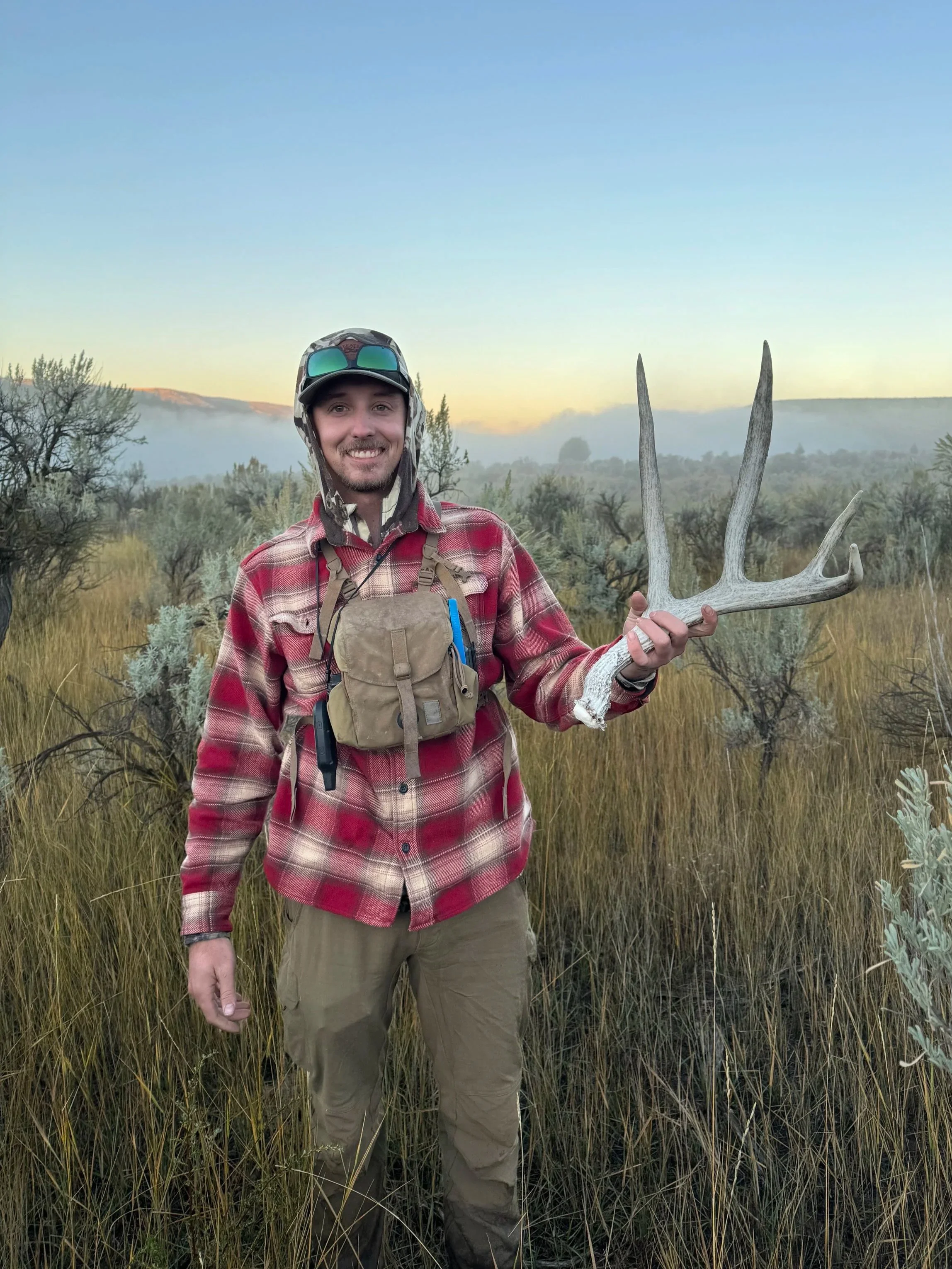 A man smiling in a wilderness area, holding a large antler, with mountains and a misty sky in the background.