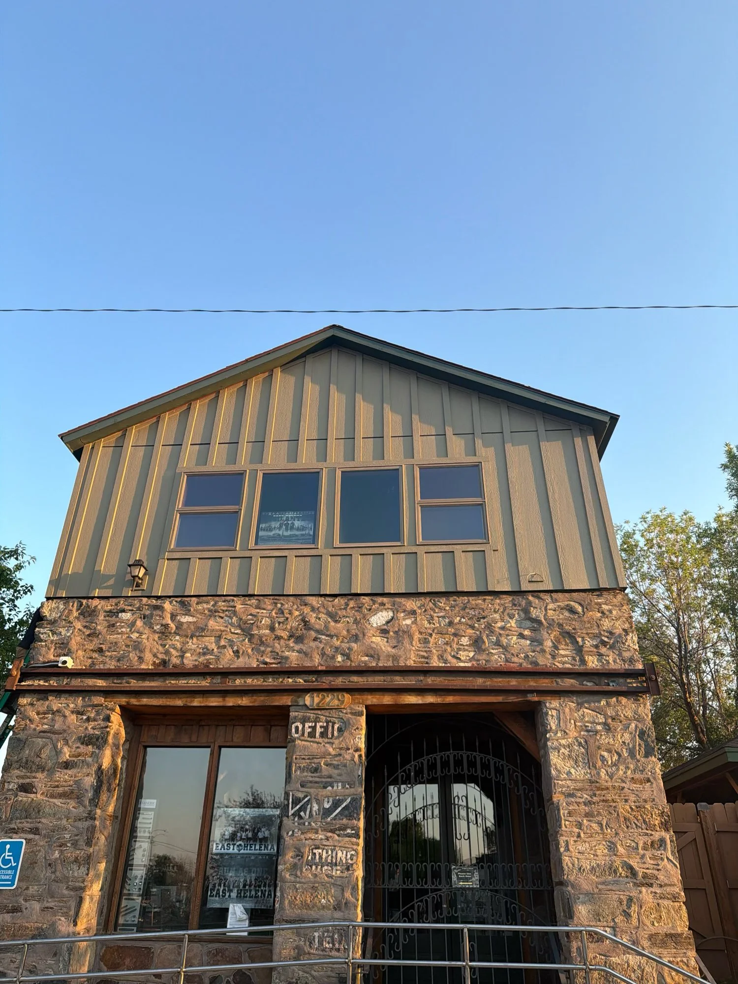 A two-story building with a stone facade and vertical wooden siding painted in a muted green color. The upper floor has three windows, and the lower floor has a large window and a wrought iron gate entrance with a sign that reads 'OFFICE.' The buildi
