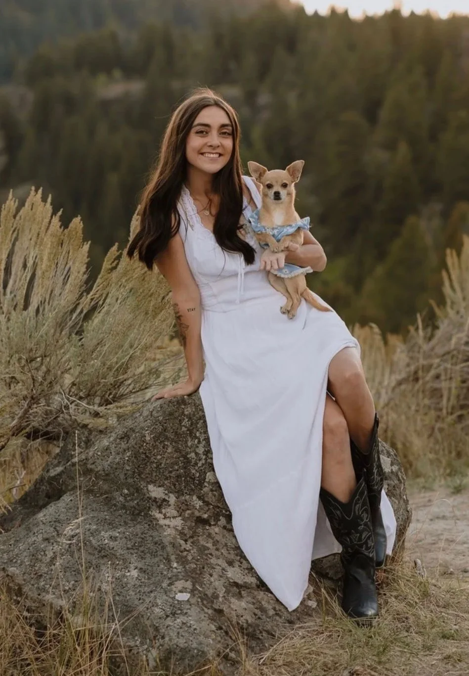 A smiling woman with long dark hair, wearing a white dress, sitting on a rock outdoors near a body of water, holding a small dog in a blue dress.