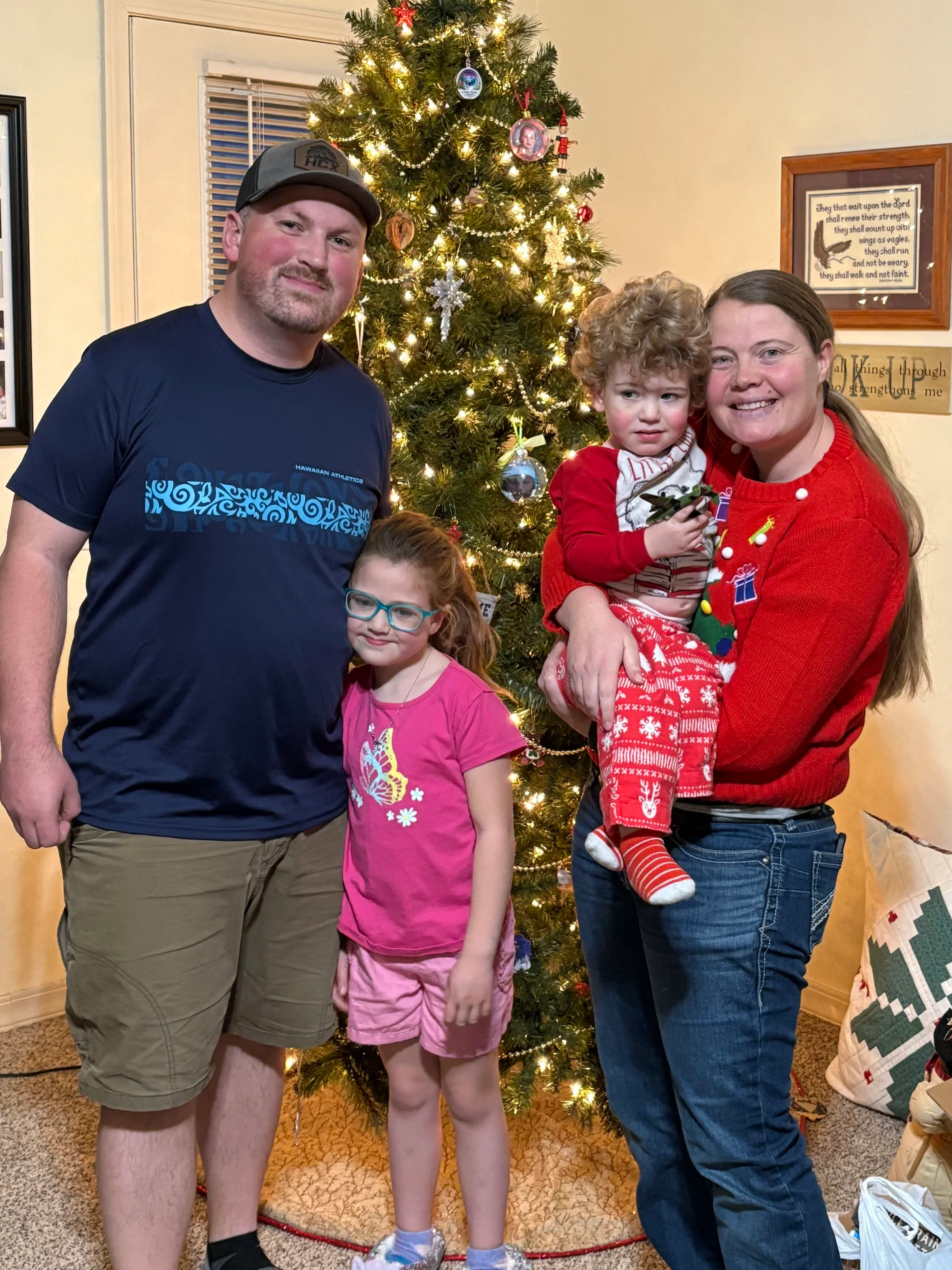 A family of four posing in front of a decorated Christmas tree. The man wears a dark blue Hawaiian shirt and khaki shorts, the woman wears a red Christmas sweater and jeans, the girl has glasses and a pink dress, and the boy is held by the woman wearing Christmas pajamas.