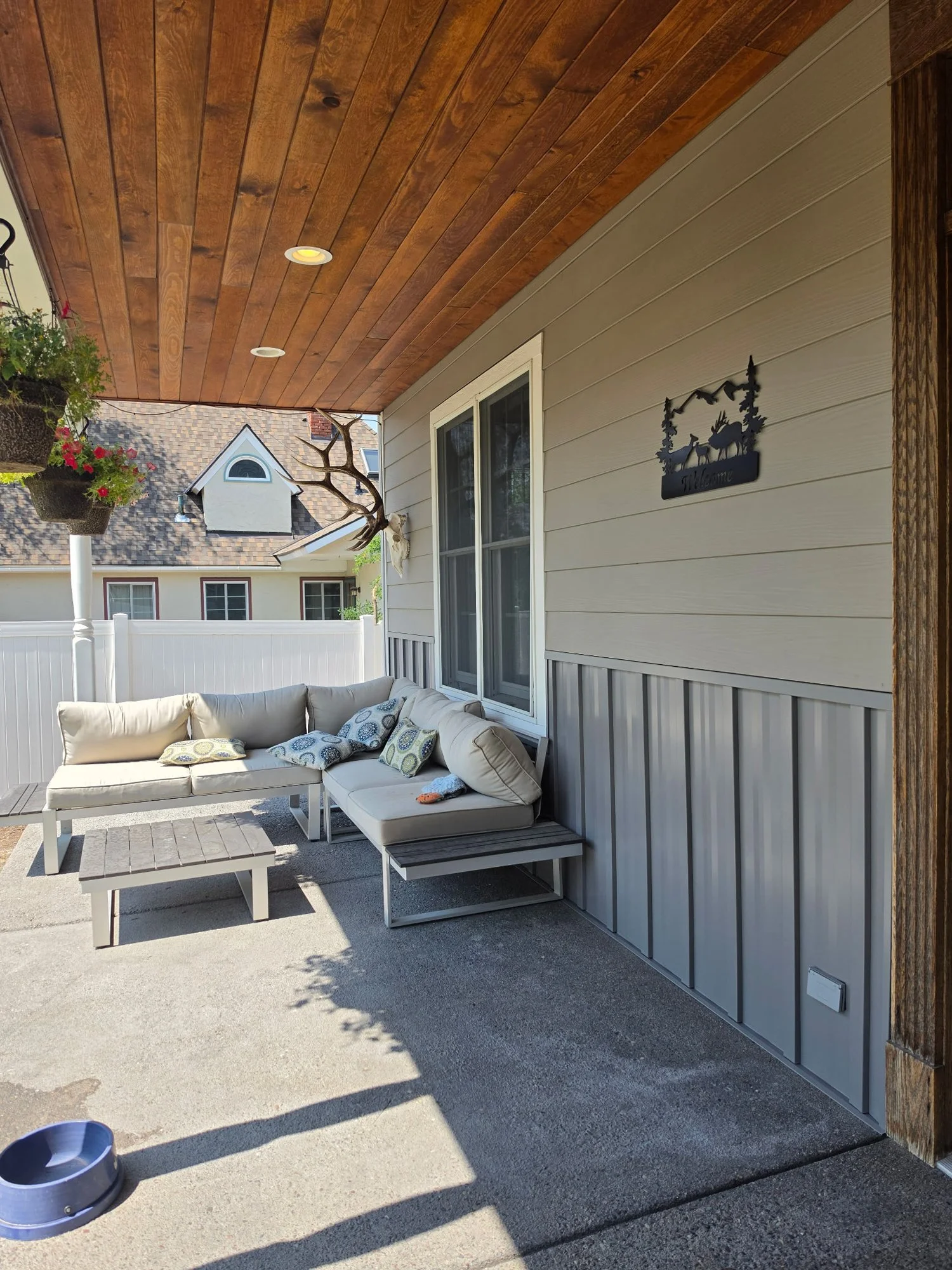 A backyard patio with a beige outdoor sofa with pillows and a matching coffee table. There are hanging planters with flowers, a dog skull mounted on the wall, and a black metal decorative sign with deer and forest motif. The porch has a wooden ceilin