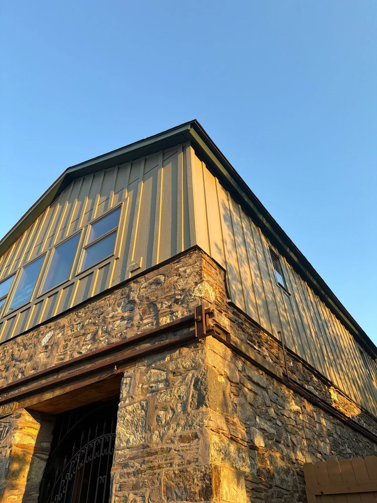 Close-up of a building's exterior corner showing stone base and upper wooden siding with vertical panels, with a clear blue sky in the background.