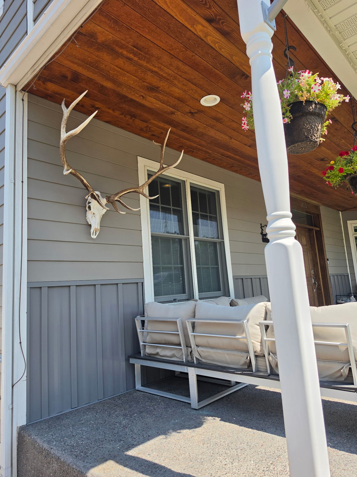 A porch area with a beige cushioned outdoor sofa and a white metal frame, situated on a concrete patio. There is a window on the house exterior, with beige siding and a darker siding on the lower part. A mount with antlers and a skull decoration is o