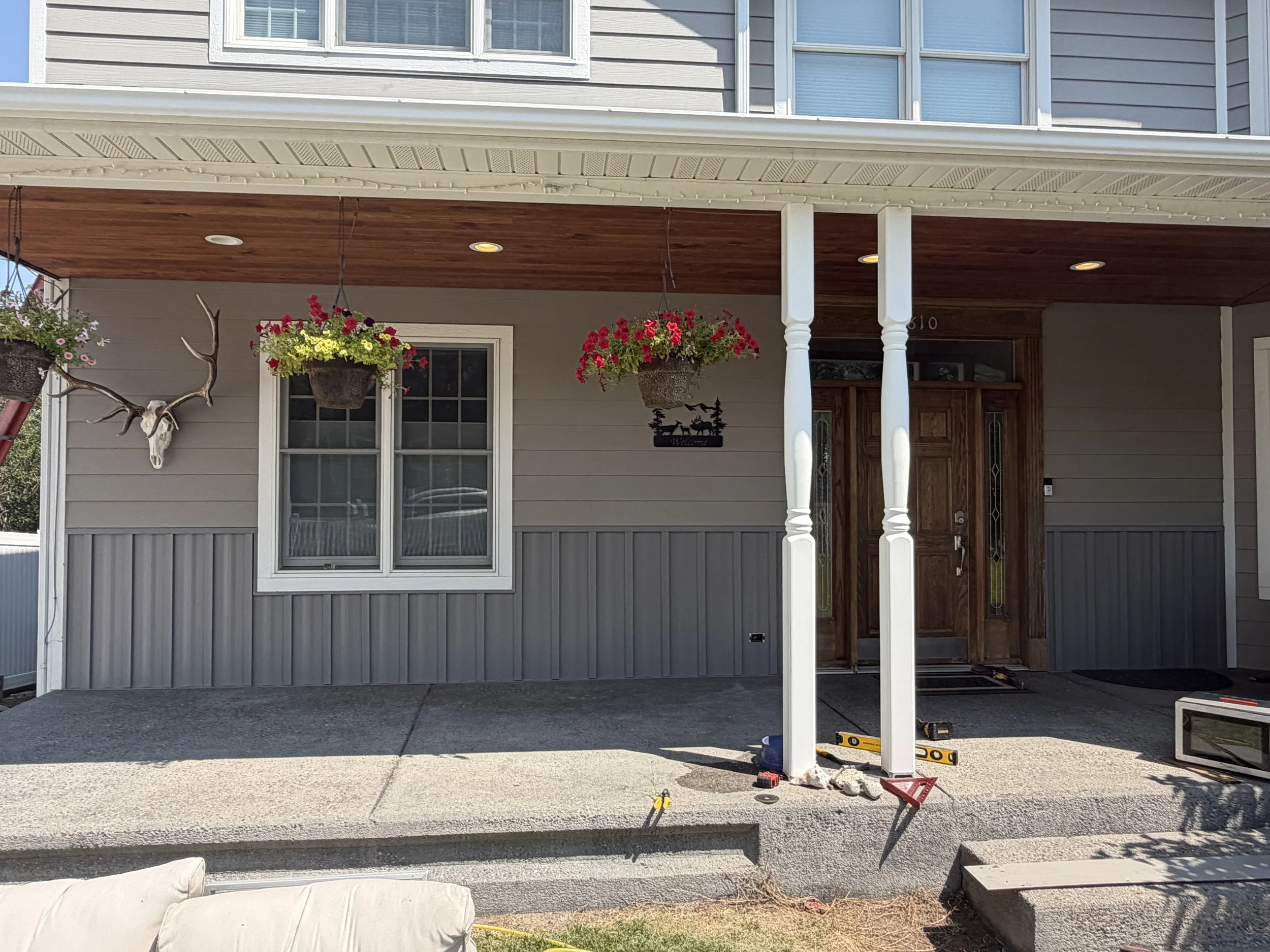 Construction site in front of a house with a covered porch, hanging flower baskets, and tools on the ground, indicating ongoing work on the porch or steps.