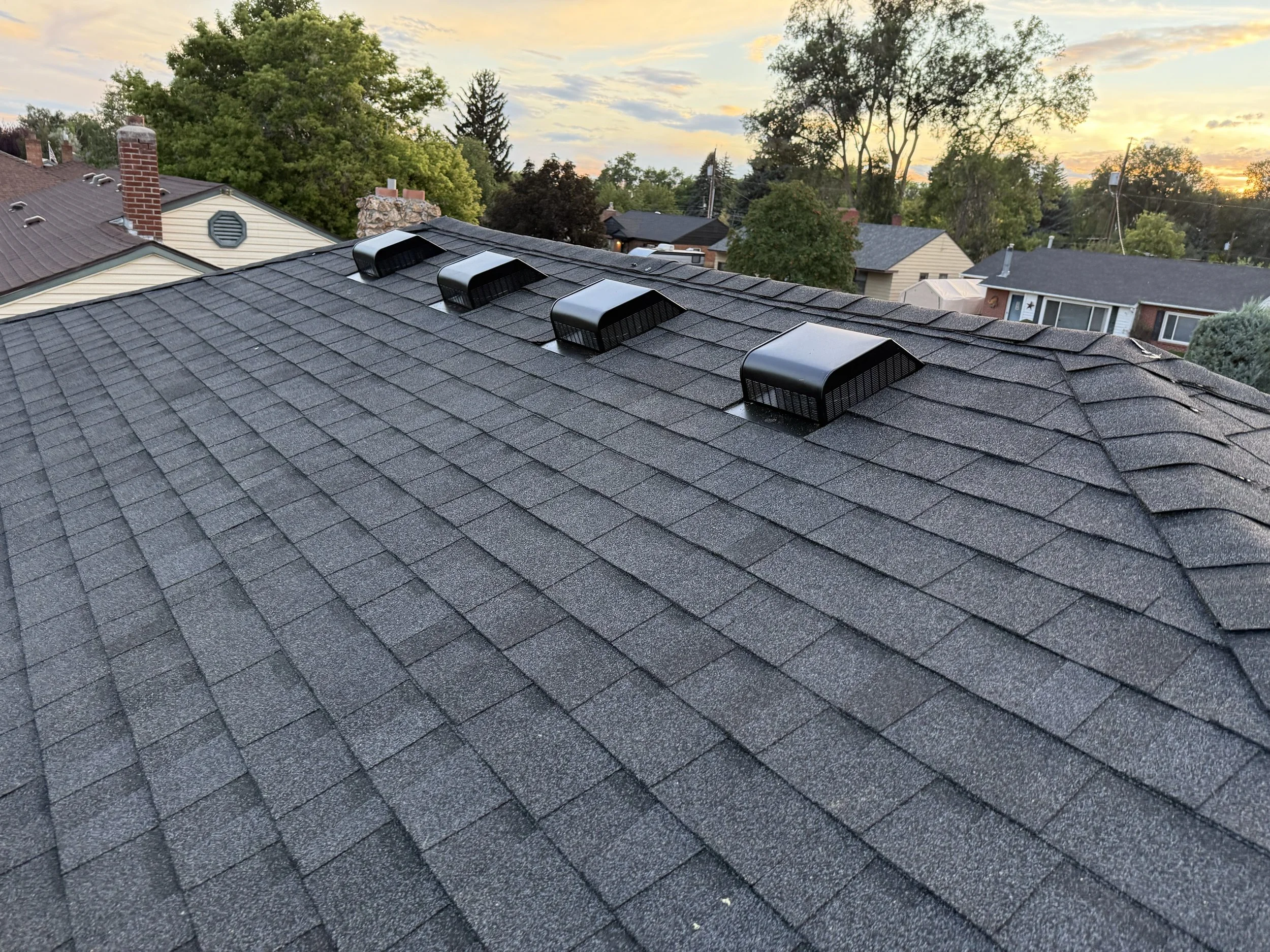A rooftop with four black vent covers on gray shingles, with trees and houses in the background during sunset.