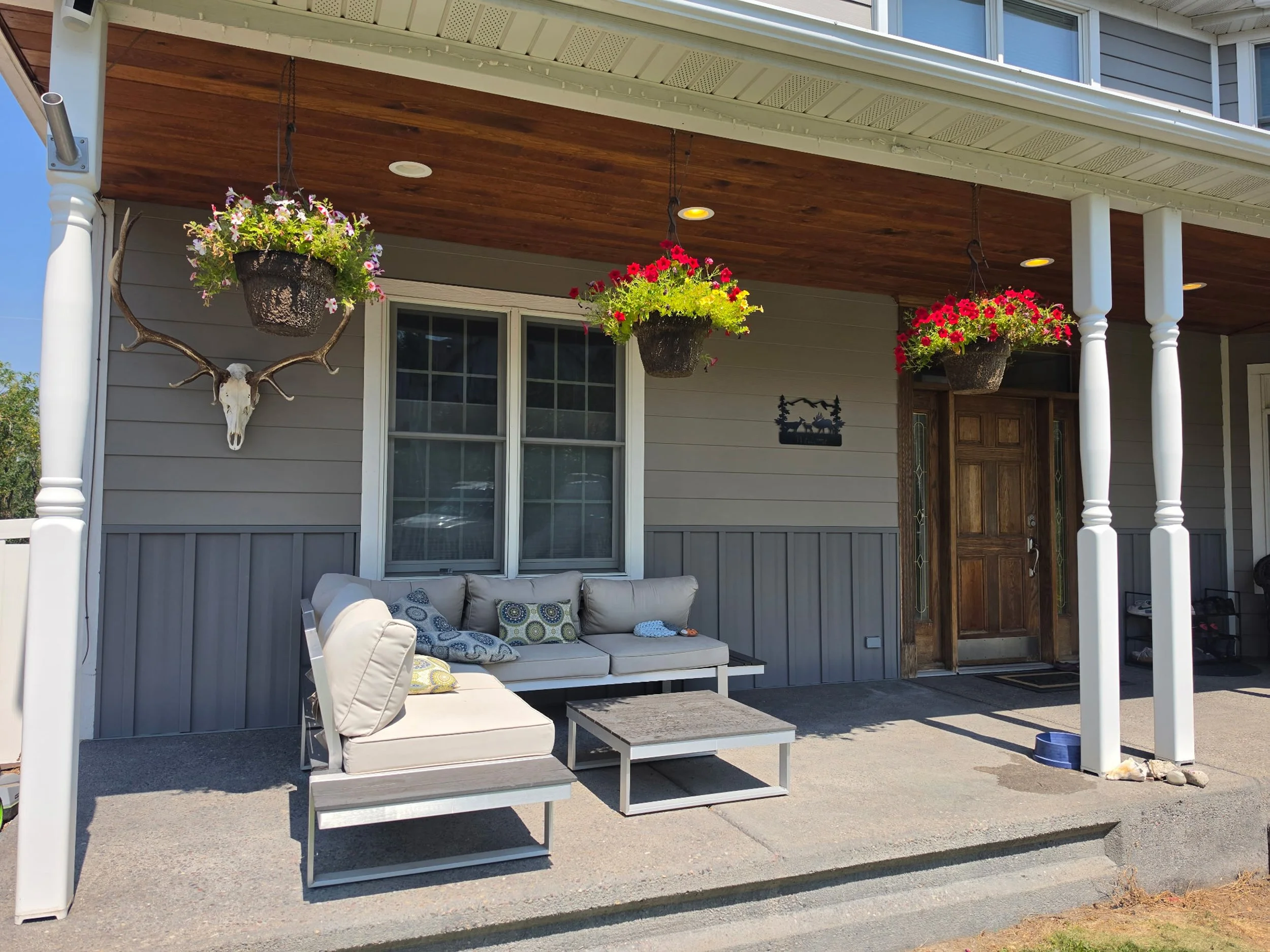 A house front porch with hanging flower pots, a deer skull with antlers, a shaded seating area with beige cushions, and outdoor furniture, with a wooden ceiling and white columns.