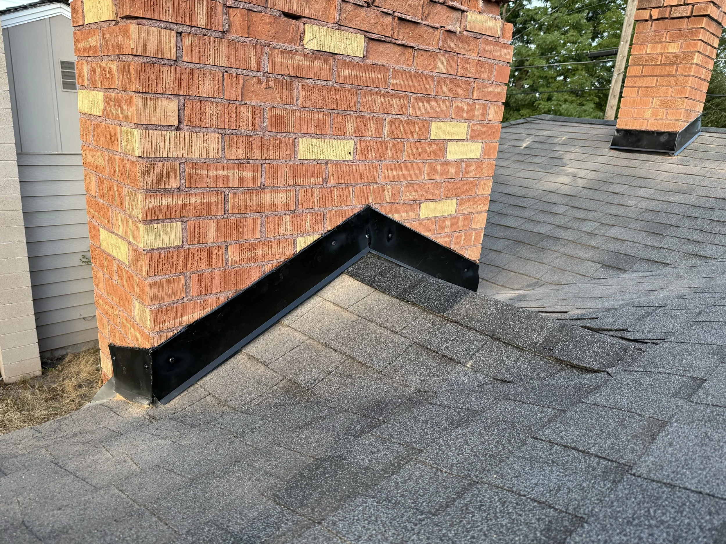Close-up of a brick chimney on a house with asphalt shingle roof and black metal flashing along the edge.
