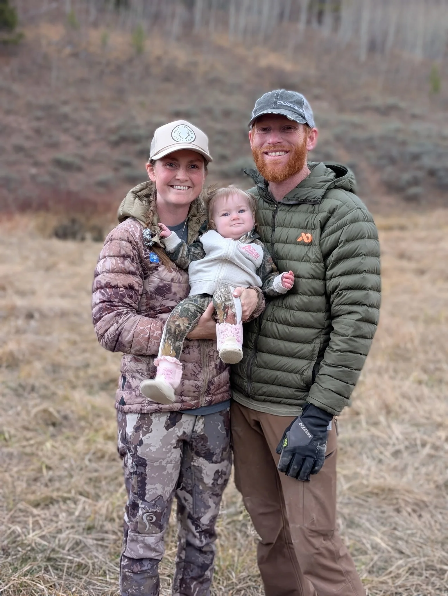 A family of three, including a woman, a man, and a young girl, smiling and posing outdoors in a natural setting with dry grass and trees in the background.