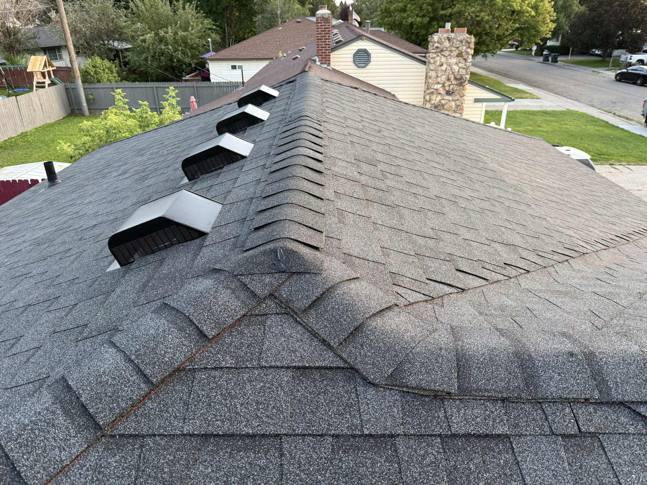 A house roof with asphalt shingles and four solar attic vents, with a residential neighborhood and trees in the background.