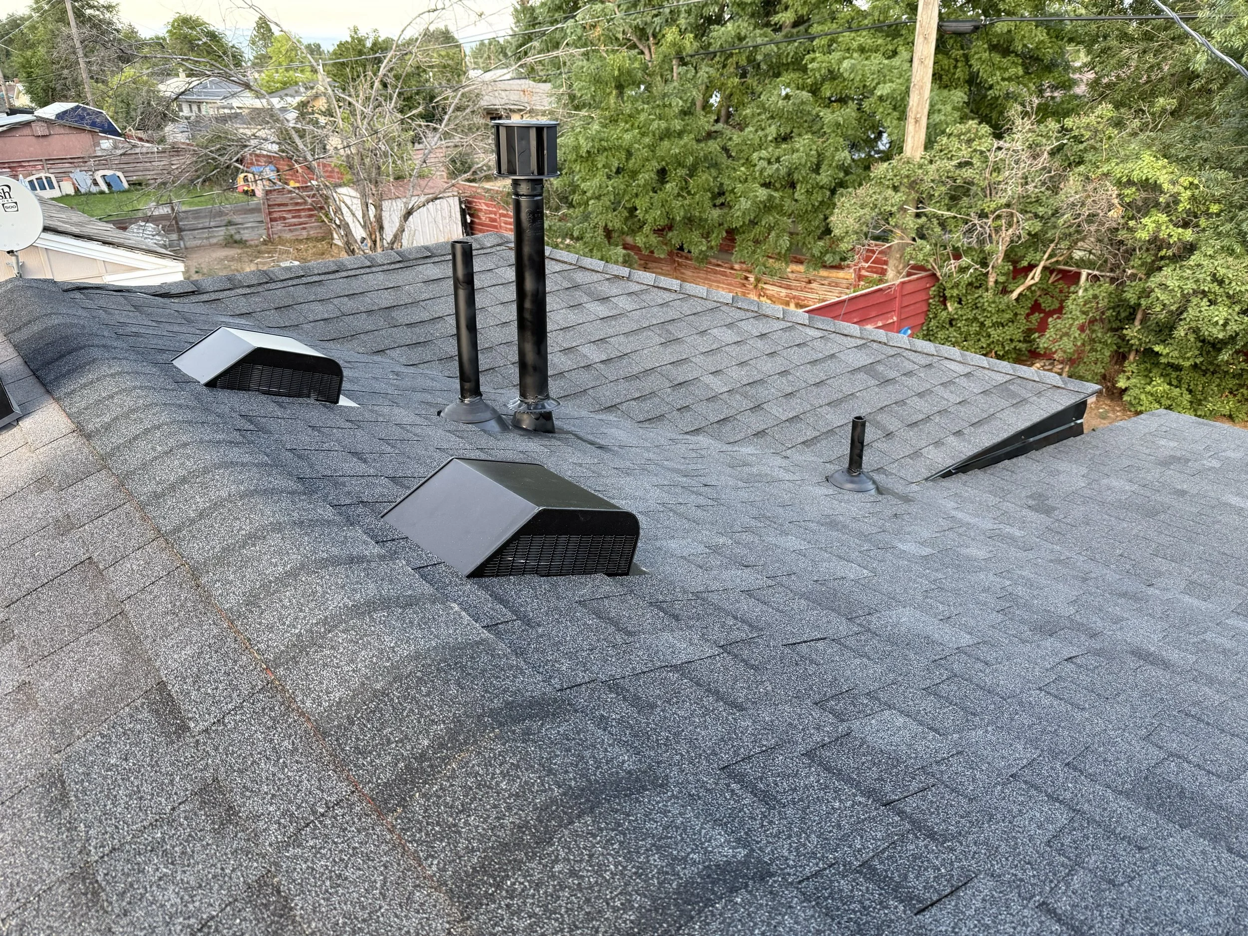 View of rooftops, trees, and power lines from a house's roof, with vents and chimney pipes visible in the foreground.