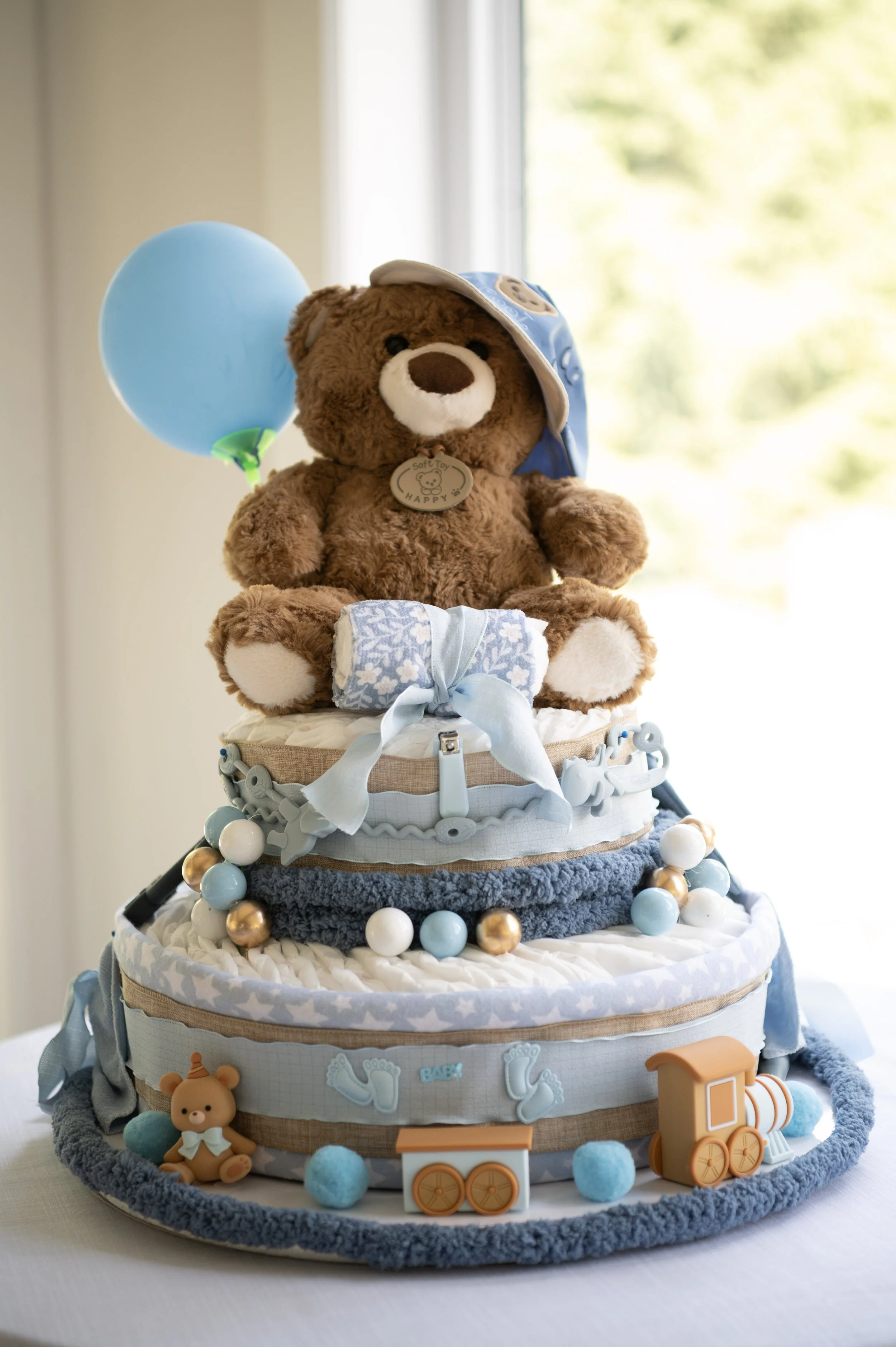 Baby shower diaper cake decorated with plush teddy bear, blue balloon, wooden toy train, plush bear, and baby-themed accessories in blue, white, and beige colors.