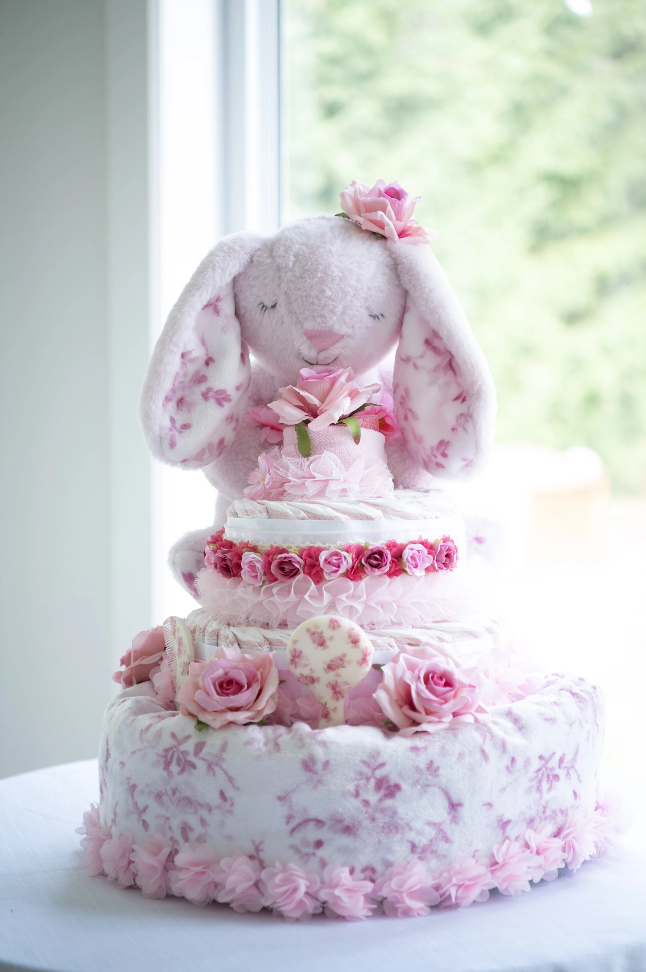 A pink and white tiered cake decorated with pink roses and floral accents, topped with a plush bunny toy with closed eyes, long ears with floral lining, and a pink flower on its head.
