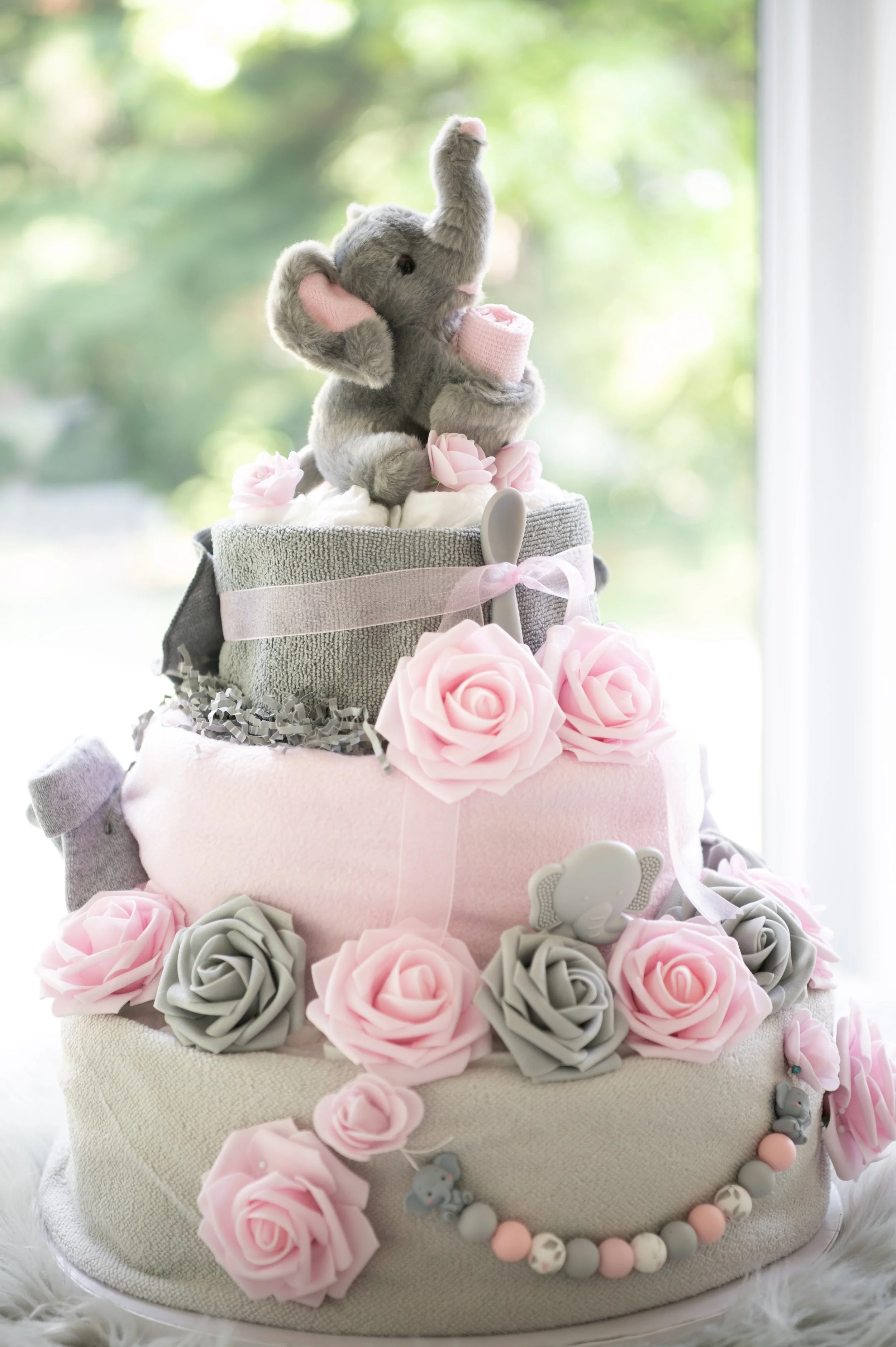 Multilayer diaper cake decorated with soft pink and gray artificial roses, a pink and gray beaded necklace, and a small plush elephant toy.