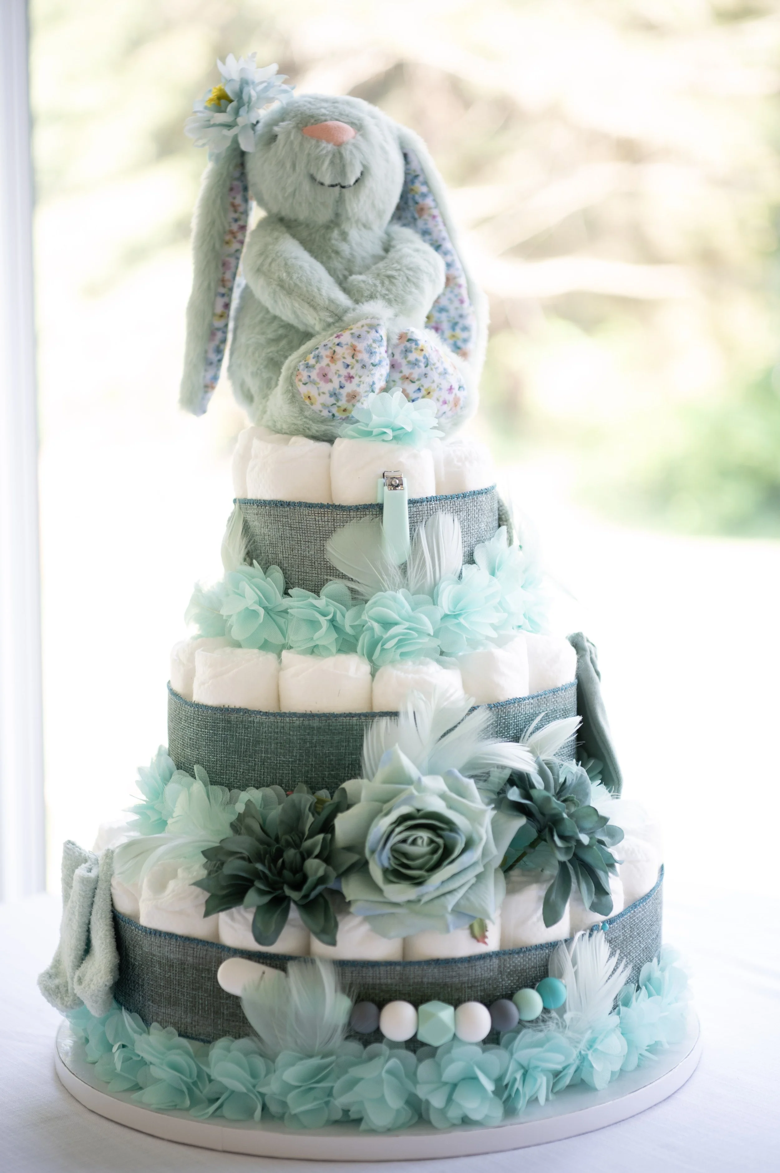 Decorative diaper cake with a plush bunny on top, adorned with flowers and pastel-colored accents, surrounded by flowers at the base.