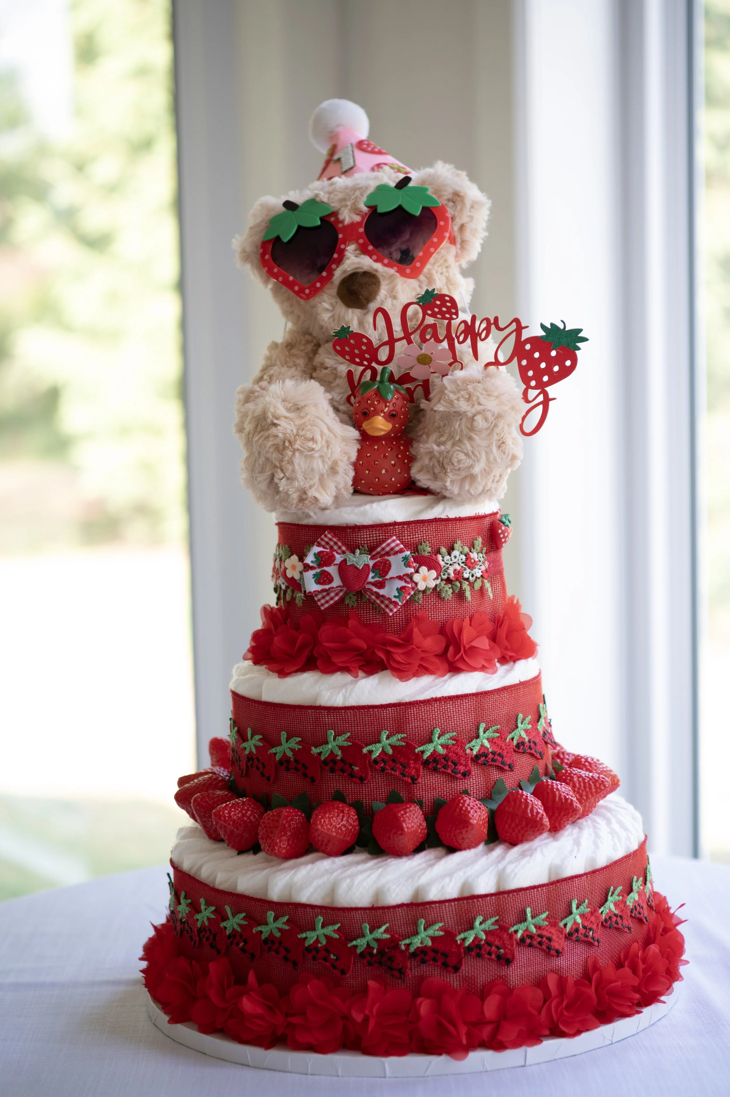 A layered strawberry-themed cake with decorative red and green strawberry patterns, strawberries on top, and a teddy bear wearing festival sunglasses and a party hat, holding a sign that says "Happy".