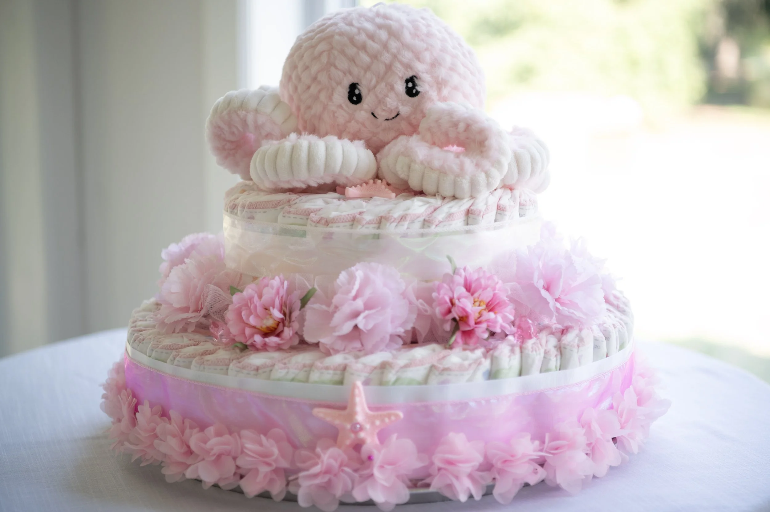A two-tier pink and white diaper cake decorated with roses, a pink star, and a plush octopus toy with a smiling face.