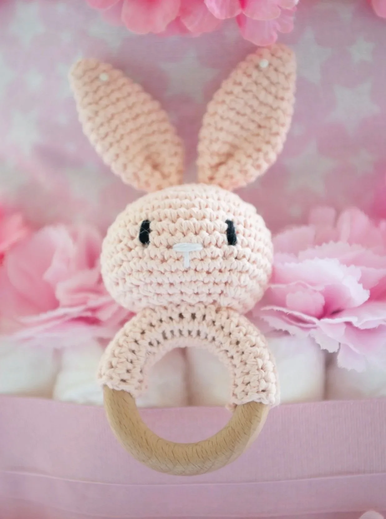 A crocheted plush bunny with pink ears, black eyes, a small white nose, and a looped wooden ring attached, set against a pink background with pink flowers and a pink box.
