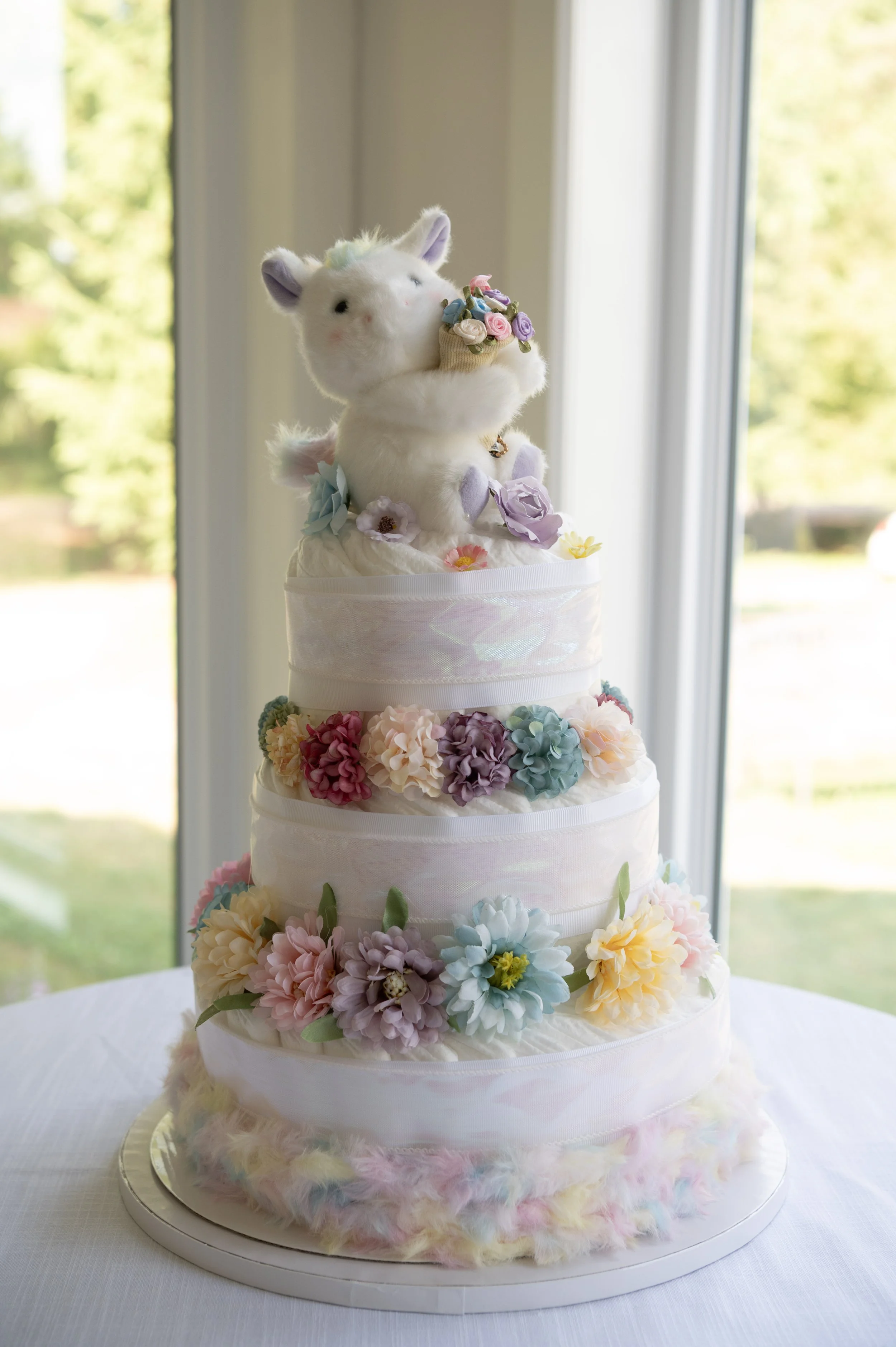 A multi-tiered cake decorated with pastel-colored flowers and a plush unicorn holding a bouquet of flowers on top.