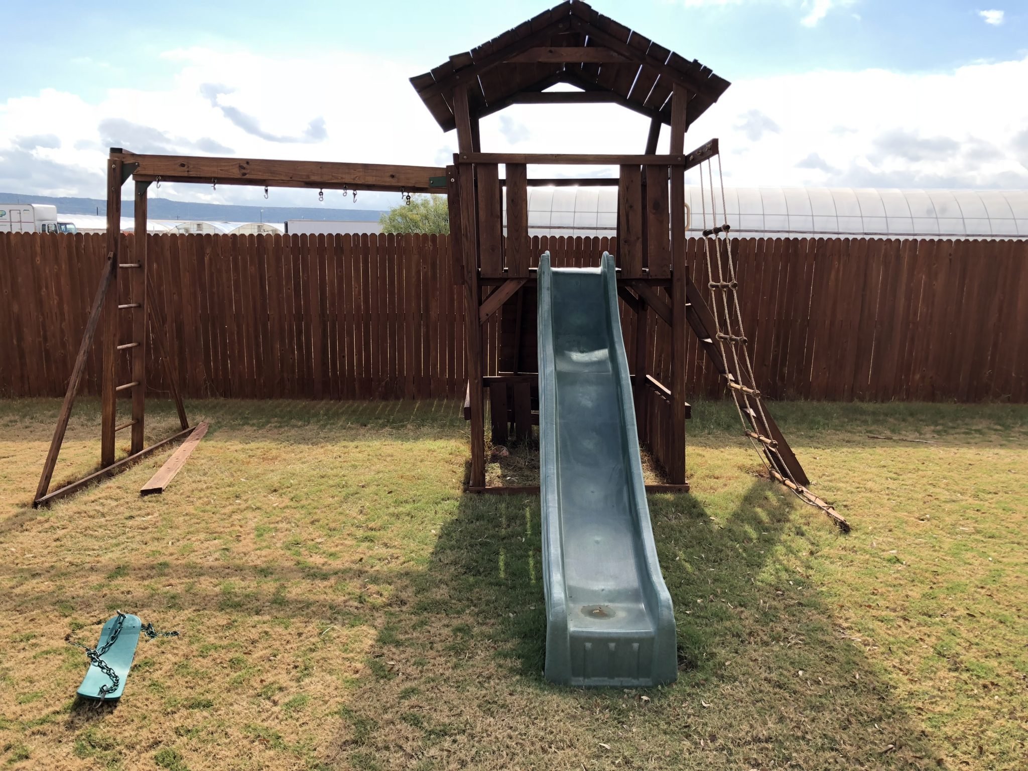 A backyard playground with a wooden play structure, slide, swings, and climbing ropes, enclosed by a tall wooden fence. There is a sunny sky with scattered clouds.
