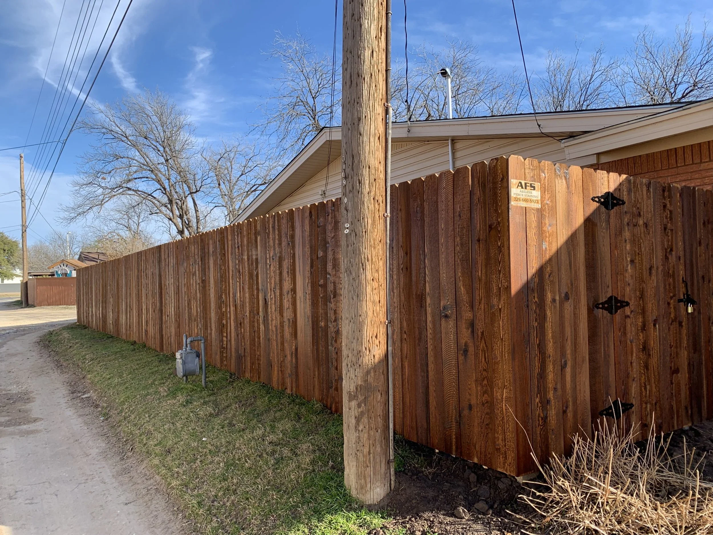 A tall wooden utility pole next to a wooden privacy fence alongside a dirt sidewalk. The sky is clear with some wispy clouds, and there are leafless trees in the background. Power lines run from the pole across the sky.