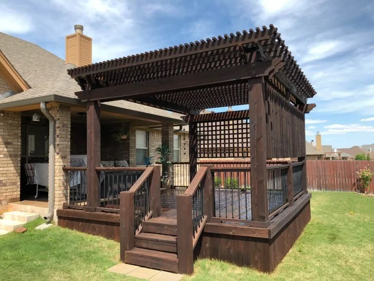 A wooden pergola on a raised deck attached to a brick house with a porch.