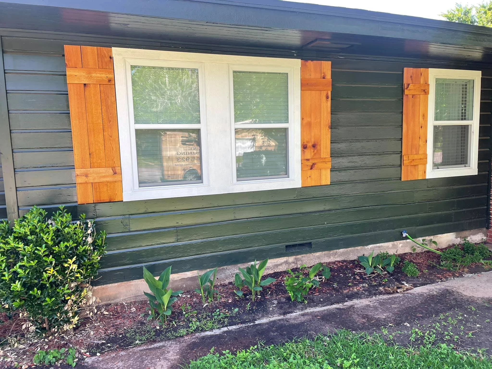 A house with dark green horizontal siding, white-framed windows, and wooden shutters. There is a garden with green plants and a bush in front of the house.