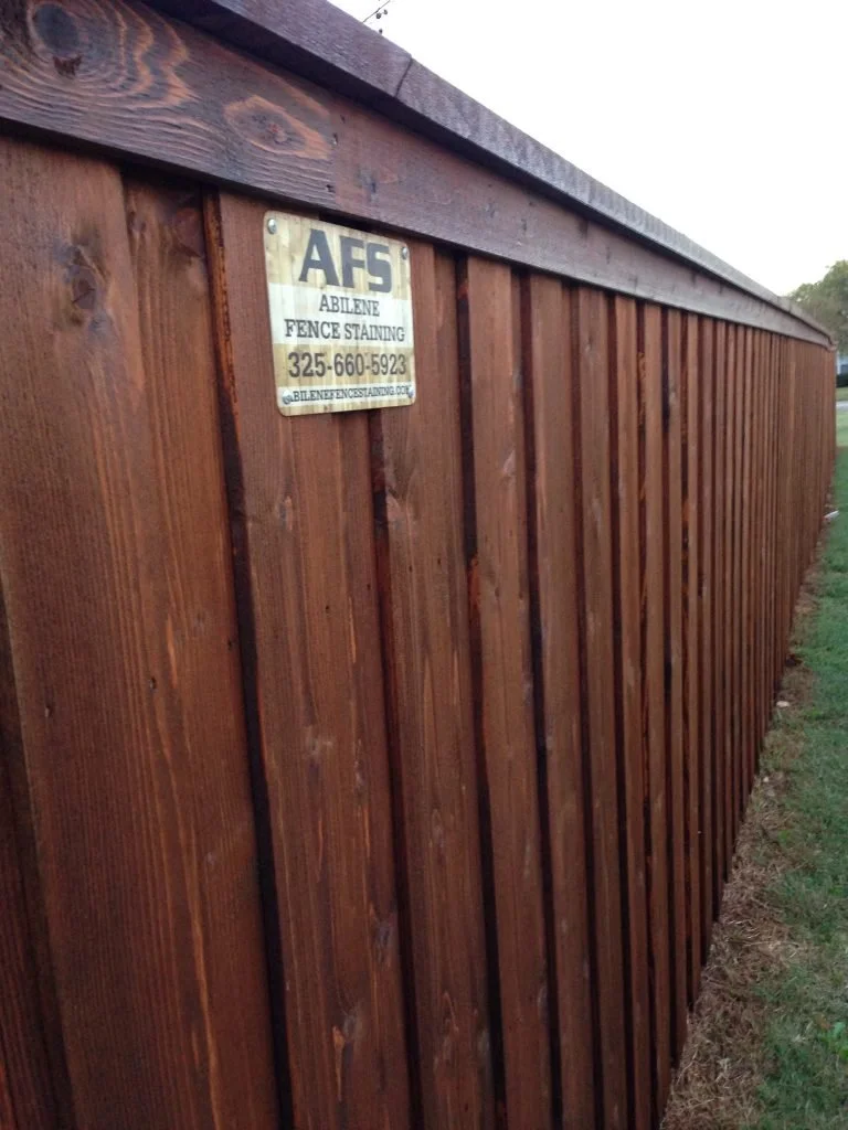 Long wooden fence with a sign that reads 'AFS Abilene Fence Staining' and a phone number, located outdoors next to a grassy area.
