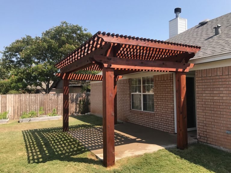 A newly built wooden pergola attached to the back of a brick house in a backyard, casting a shadow on the patio below.
