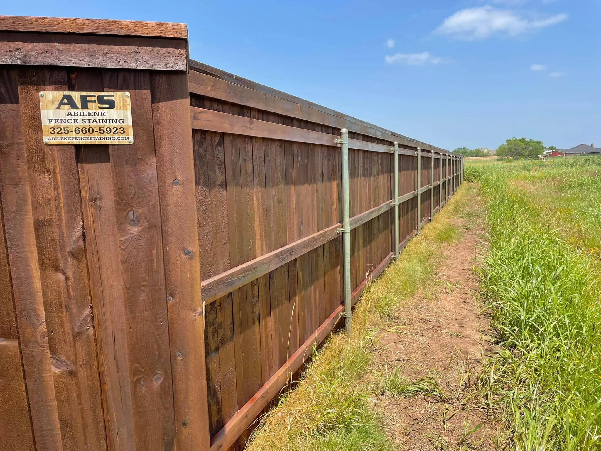 A tall wooden fence extending into the distance, with metal posts and rails, in a grassy area under a clear blue sky with a few clouds.