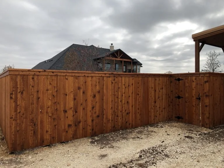 New wooden fence in backyard with gravel ground and a large house with a porch in the background under cloudy sky.