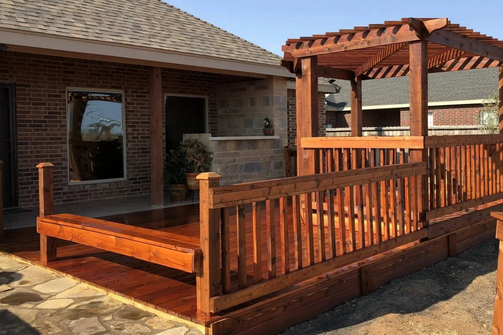 A backyard deck with a wooden pergola, surrounded by a wooden railing, attached to a brick house with stone accents.